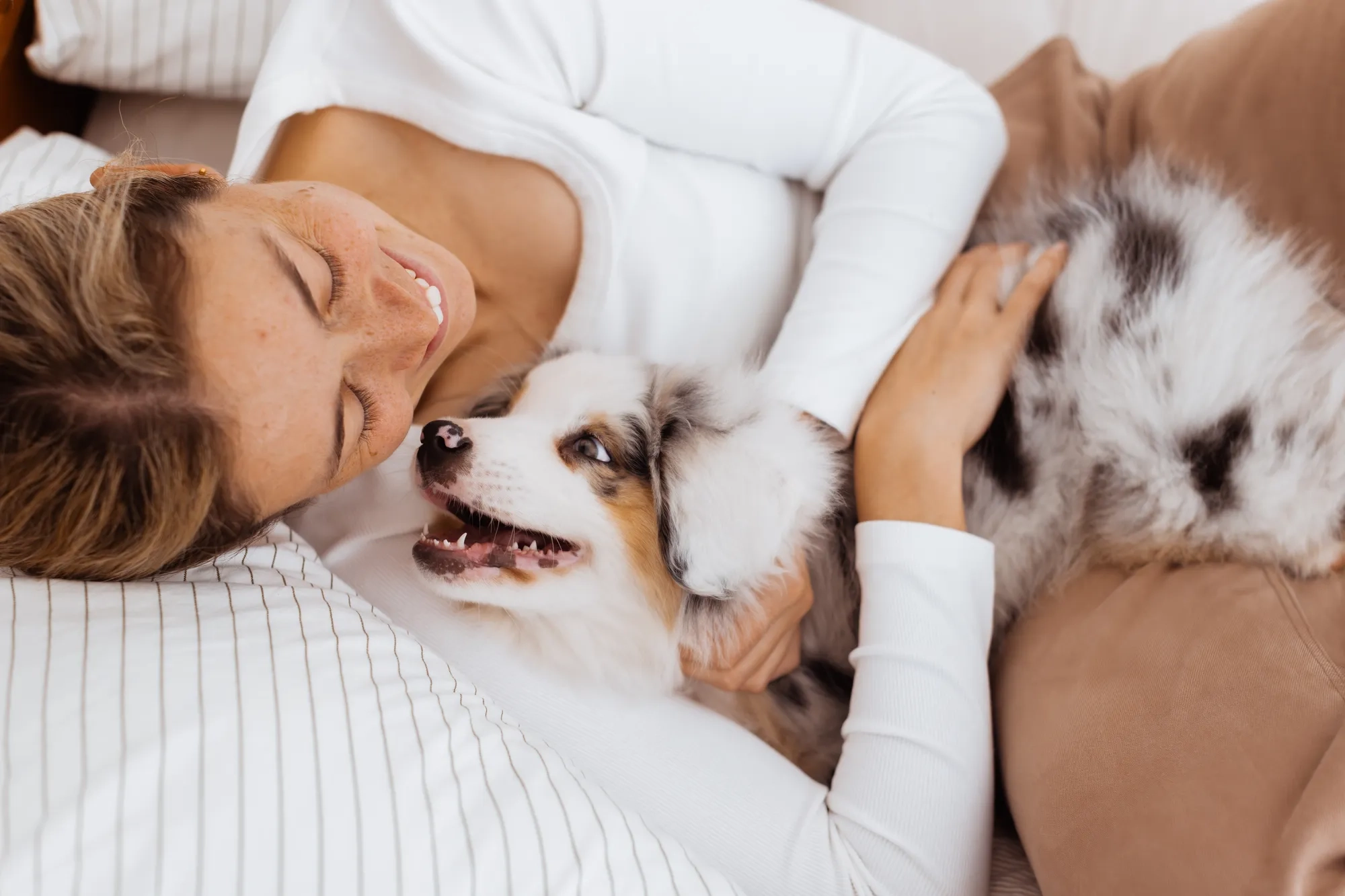A woman lying on a bed in a white long-sleeve shirt smiles while cuddling her playful Australian shepherd puppy, capturing the essence of professional puppy photos. 